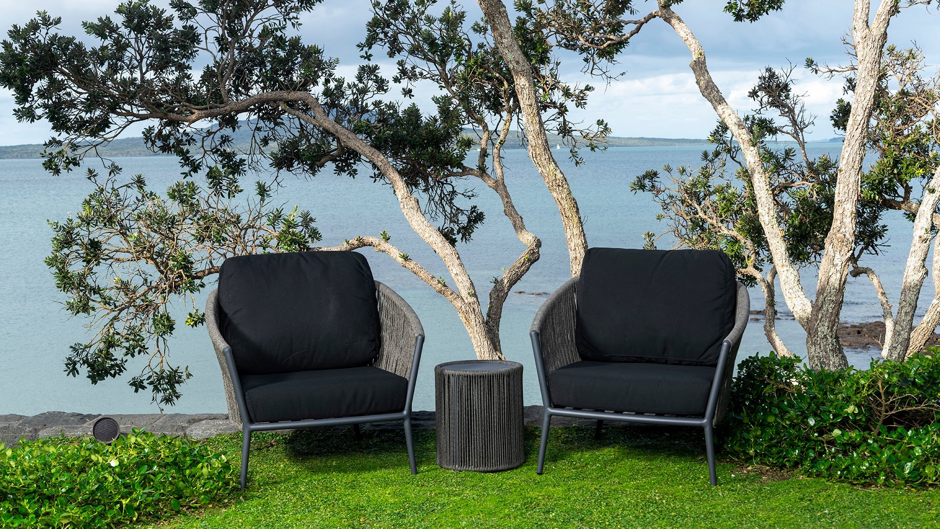 Two Washington outdoor club chairs with black cushions set against a scenic beach backdrop, flanked by lush greenery and overlooking calm sea waters.