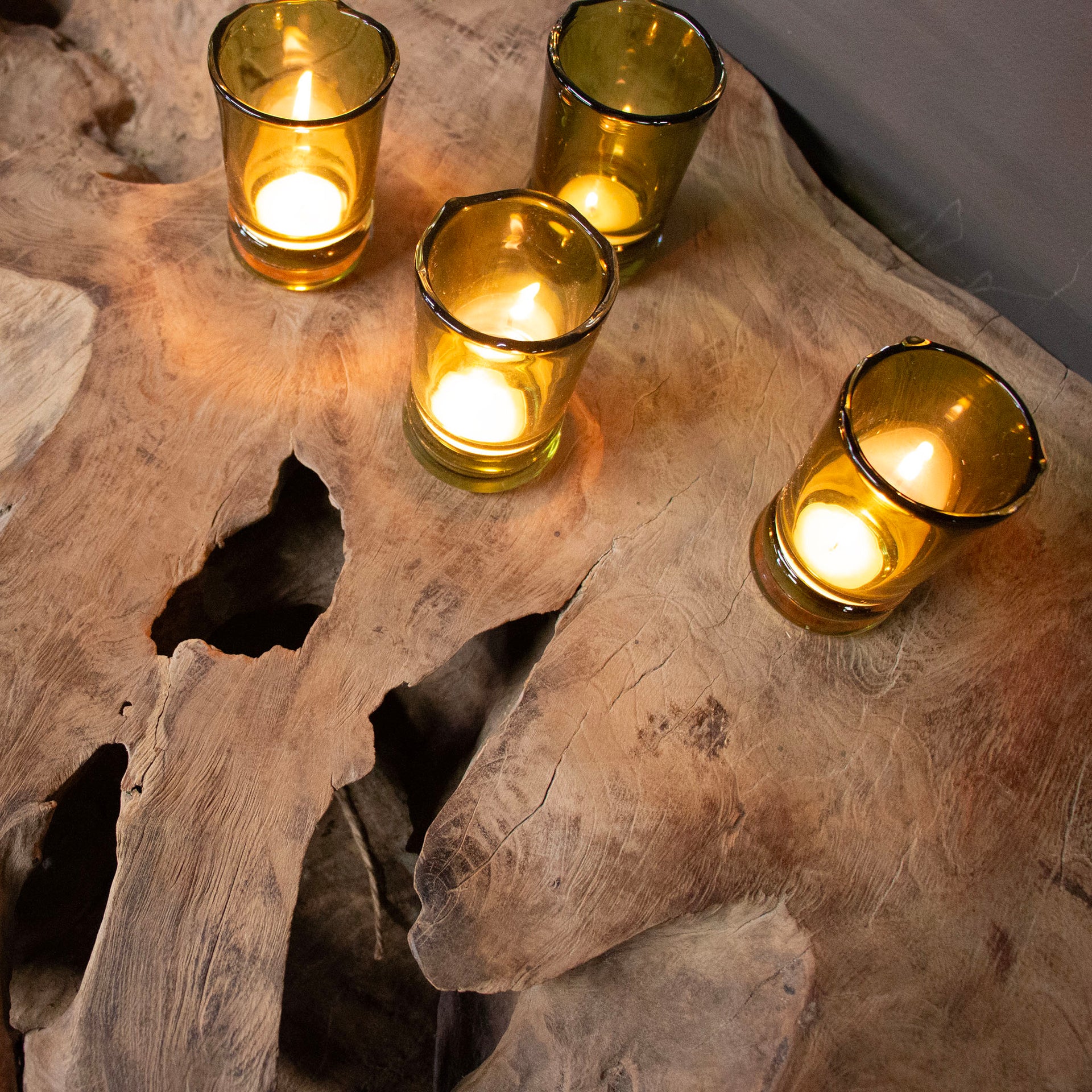 Four glass candles holders on top of a teak wood console table with candles burning.